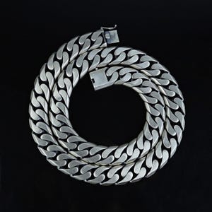 May include: A silver curb chain necklace with a substantial weight. The necklace is made up of thick, interlocking links and has a secure clasp. The chain is shown against a black backdrop, emphasizing its metallic shine and striking design.