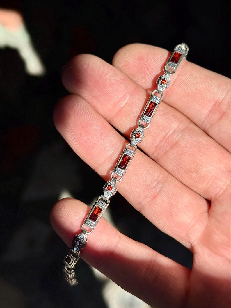 May include: A silver bracelet with rectangular and diamond-shaped red gemstones. The bracelet is held in a hand, with the sunlight reflecting off the polished metal and stones. The bracelet is a piece of jewelry.