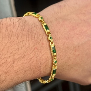 May include: Gold-toned bracelet featuring rectangular emerald-green stones and smaller round green stones. The bracelet has an ornate design with decorative elements. The jewelry is displayed on a person's arm.