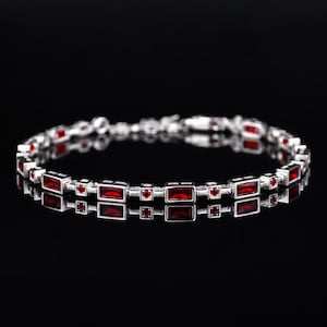 May include: A silver-toned bracelet featuring a repeating pattern of rectangular and circular red gemstones. The design alternates between elongated baguette-cut red stones and small round red stones set within a metallic frame structure.