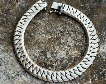 May include: A silver-toned bracelet with a geometric, zig-zag design. The bracelet is open, showing the clasp mechanism. The links are triangular and create a textured surface. The bracelet is likely made of metal and is displayed on a textured surface.