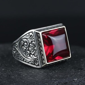 Peut inclure: Bague en argent avec une grosse pierre rouge carrée. La monture présente des motifs floraux et des volutes complexes. La pierre est sertie dans un cadre carré avec un motif tressé. La bague est présentée sur une surface sombre.