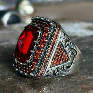 Puede incluir: Un anillo de plata con una gran piedra roja rodeada de piedras rojas más pequeñas. El anillo tiene detalles intrincados y ornamentados.