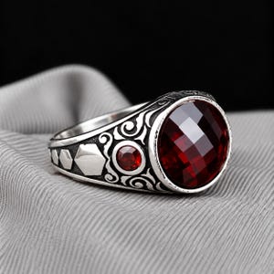 Puede incluir: Un anillo de plata con una gran piedra preciosa redonda y facetada de color rojo intenso como pieza central. La banda está decorada con volutas intrincadas, una piedra roja redonda más pequeña y formas hexagonales geométricas.
