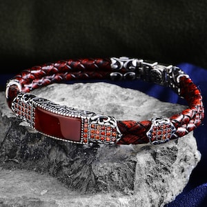 Può includere: Un bracciale in pelle intrecciata con un elemento centrale rettangolare in pietra rossa e dettagli argentati. Il bracciale ha una chiusura a fibbia ed è esposto su una superficie di pietra grigia. La pelle è di colore rosso scuro.