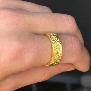 Puede incluir: Un anillo dorado con un intrincado patrón floral grabado alrededor de la banda. El anillo se muestra en un dedo, mostrando el diseño detallado y el acabado pulido del metal. El anillo es una pieza de joyería.