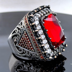 May include: A silver ring featuring a large red gemstone surrounded by smaller white gemstones. The ring has intricate filigree details and is set in a square shape.