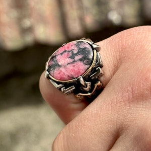 May include: A silver ring with a pink and black gemstone set in a detailed silver and gold setting.