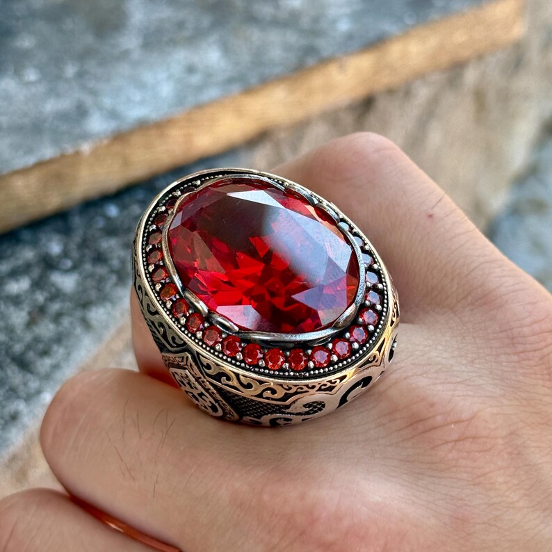 Puede incluir: Un anillo de plata ornamentado con una gran gema roja ovalada. El anillo est&aacute; rodeado de piedras rojas m&aacute;s peque&ntilde;as y detalles de filigrana, creando un dise&ntilde;o lujoso y llamativo.