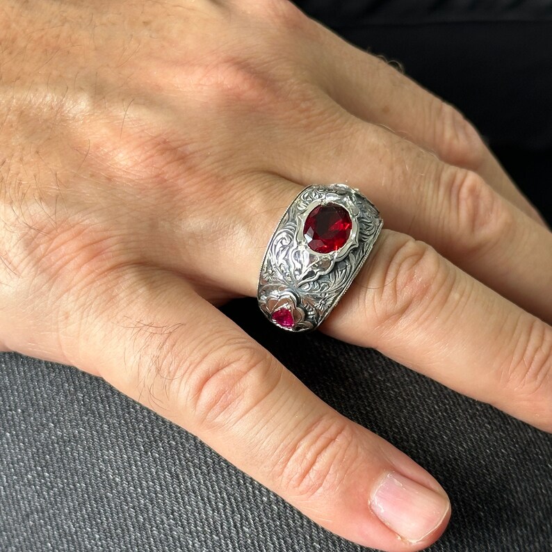 Peut inclure: Une bague en argent avec un motif orn&eacute; et une grosse pierre pr&eacute;cieuse rouge. La bague est port&eacute;e sur un doigt.