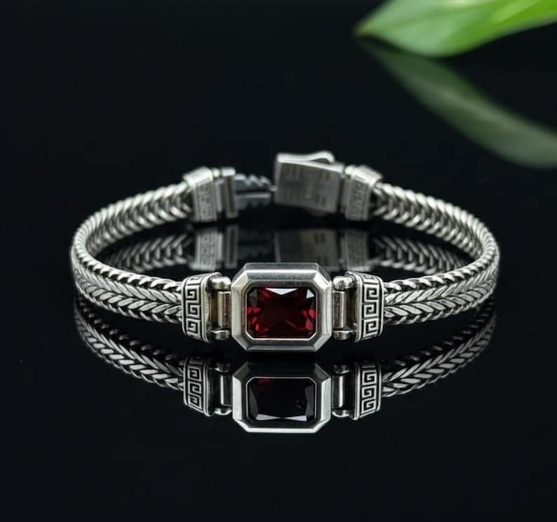May include: A silver bracelet with a woven chain design and a rectangular, deep red gemstone at the center. The bracelet has a secure clasp and decorative geometric patterns. The jewelry is displayed on a reflective black surface.
