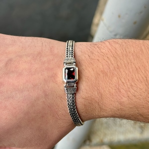 May include: Silver bracelet featuring a rectangular, deep red gemstone at the center. The bracelet has a woven chain design with decorative rectangular links on either side of the gemstone. The bracelet is worn on a wrist.