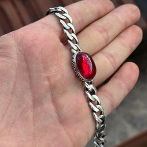 Könnte beinhalten: Ein silbernes Armband mit einem großen, ovalen, roten Edelstein, der in einer dekorativen Lünette gefasst ist. Das Armband hat ein dickes Panzerketten-Design. Der Edelstein ist der Blickfang und setzt einen Farbtupfer auf die Silberkette.