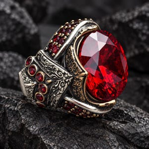 May include: A silver ring featuring a large, oval-cut, deep red faceted gemstone as the centrepiece. The band is decorated with intricate, dark-toned metal engravings and is accented with multiple small, round, deep red stones along the sides.
