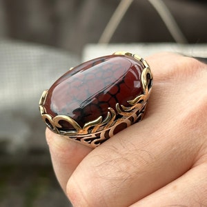 Peut inclure: Une bague ornée d'une grosse pierre ovale. La pierre est un brun-rouge poli avec des veines sombres, sertie dans une monture en métal doré. La monture présente des filigranes détaillés. Un bijou saisissant.