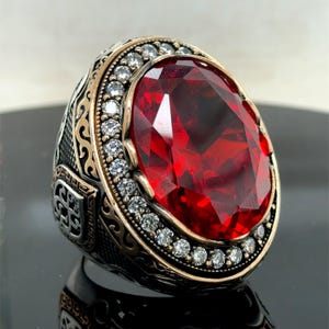 Pode incluir: Anel de prata com uma pedra preciosa vermelha oval como ponto focal. A pedra é rodeada por uma fila de pequenas pedras transparentes e uma faixa dourada. A banda do anel apresenta intrincados detalhes em metal escuro.