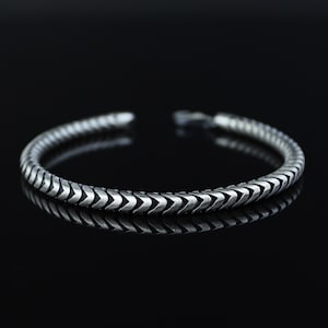 May include: A silver-colored cuff bracelet with a chevron pattern. The bracelet is open at the ends and reflects on a dark surface. The design features a series of repeating arrow-like shapes.