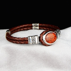 May include: A brown leather bracelet with a silver metal clasp and a red oval stone. The clasp has a Greek key design.