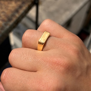 May include: A polished gold-tone signet ring featuring a smooth, rectangular flat top surface. The band is solid and tapers slightly toward the bottom, showcasing a reflective metallic finish that catches the light across its clean, minimalist edges.