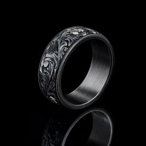 May include: A dark gray ring with intricate floral engravings. The ring features a detailed pattern of flowers and leaves, with a beaded border. The ring is set against a black background, highlighting the design.