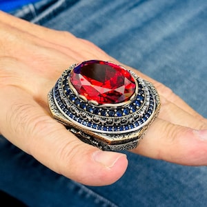 Puede incluir: Un anillo ornamentado con una gran gema roja como pieza central. El anillo está rodeado por una banda de pequeñas piedras azul oscuro y un intrincado trabajo de metal. El diseño del anillo es detallado y llamativo.