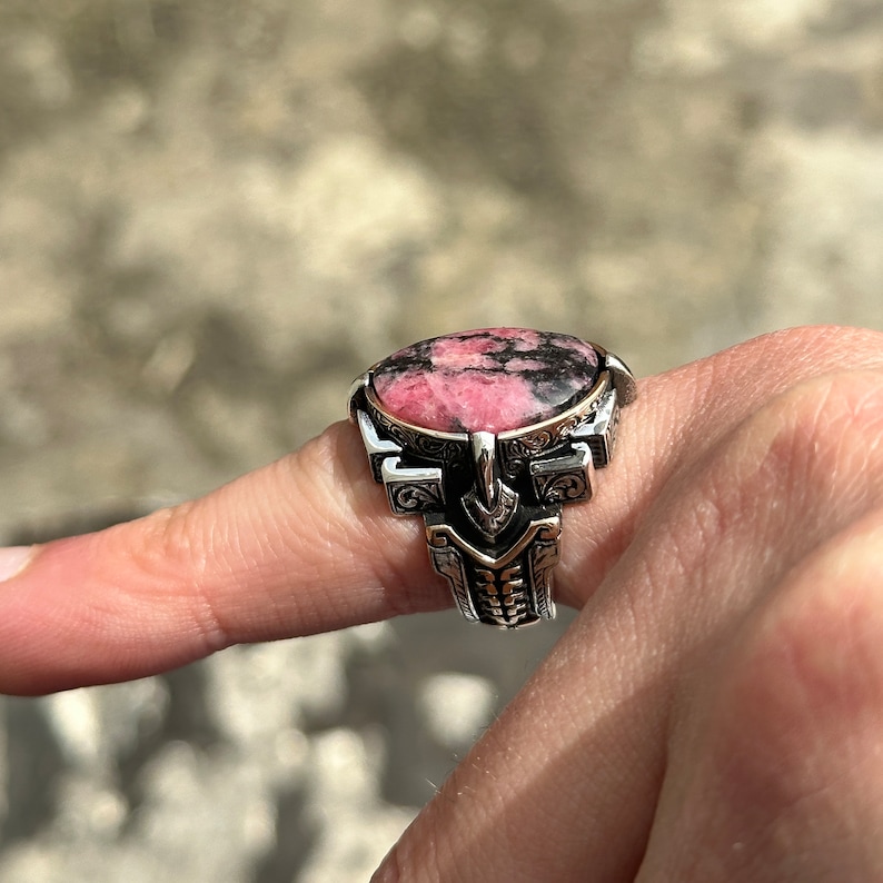 May include: A silver ring with an ornate design features a pink and black gemstone. The ring is worn on a finger.