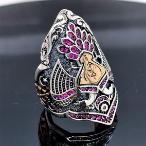 Może przedstawiać: Szczegółowy srebrny pierścionek ze złotym akcentem, z centralnym wzorem i różowymi kamieniami szlachetnymi. Pierścionek ma grawerowany wzór i ciemne tło. Projekt jest skomplikowany i szczegółowy.