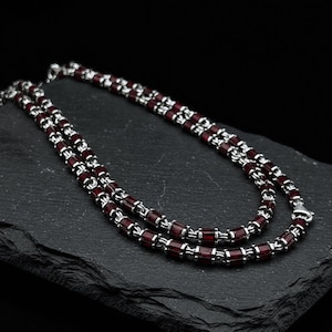 May include: A double-strand necklace featuring a chain composed of alternating silver-toned metal links and cylindrical deep red beads. The necklace is arranged in a layered loop, showcasing a consistent pattern of metallic and dark red segments.