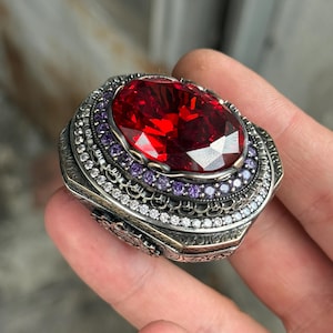 Pode incluir: Um anel de prata ornamentado com uma grande pedra vermelha de corte oval no centro. O anel é adornado com pedras roxas e brancas menores e detalhes intrincados de filigrana.