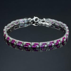 Op de afbeelding: Een zilveren armband met afwisselende rijen rechthoekige, dieproze edelstenen en kleine, heldere stenen. De armband wordt weerspiegeld in een donker, glanzend oppervlak, wat het ingewikkelde ontwerp en de sluiting benadrukt.