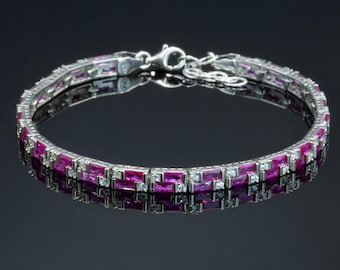 Peut inclure: Un bracelet en argent orné de rangées alternées de pierres précieuses rectangulaires rose foncé et de petites pierres claires. Le bracelet se reflète sur une surface sombre et brillante, mettant en valeur son design et son fermoir.