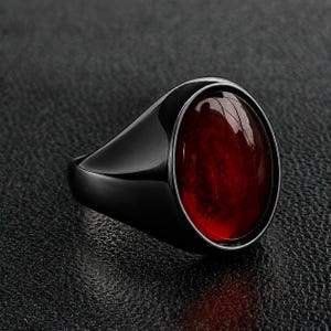 May include: A signet ring featuring a smooth, polished black metal band and a large, oval-shaped, deep red gemstone set in the centre. The stone has a glossy, reflective surface with subtle internal textures visible under the light.