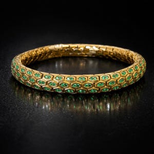 May include: A gold-toned bracelet with a honeycomb design, featuring small green gemstones set within each cell. The bracelet is displayed against a dark background, highlighting its intricate details and luxurious appearance.