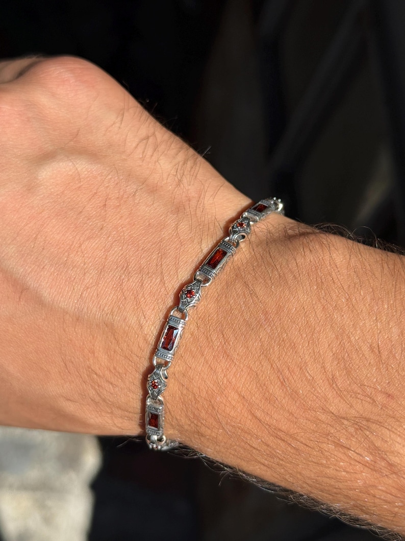 May include: Silver bracelet with rectangular and round red gemstones. The bracelet is made of silver-colored metal and features an ornate design. The gemstones are set in a row, alternating between rectangular and round shapes.