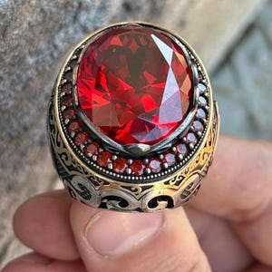 Puede incluir: Un anillo ornamentado con una gran gema roja ovalada y facetada. La banda de plata presenta intrincados filigranas doradas y peque&ntilde;as gemas rojas alrededor de la piedra principal, creando una apariencia lujosa.