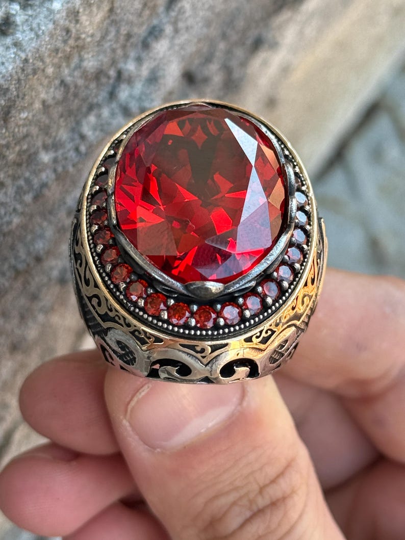 Puede incluir: Un anillo ornamentado con una gran gema roja ovalada y facetada. La banda de plata presenta intrincados filigranas doradas y peque&ntilde;as gemas rojas alrededor de la piedra principal, creando una apariencia lujosa.