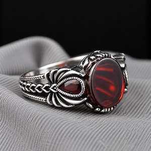 May include: A silver ring featuring a large, oval-shaped, deep red gemstone as the centrepiece, flanked by smaller, teardrop-shaped red stones. The metal band displays intricate, textured, and engraved detailing along the sides and settings.
