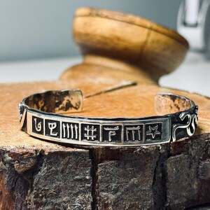 Puede incluir: Brazalete de plata con símbolos grabados y un diseño de estrella. El brazalete abierto se muestra sobre una superficie de madera. El ancho del brazalete es de aproximadamente 1,3 cm.