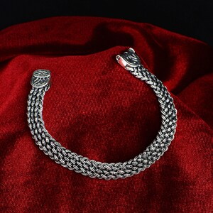 May include: A silver bracelet with a braided chain design and a clasp closure. The bracelet is on a red velvet background.