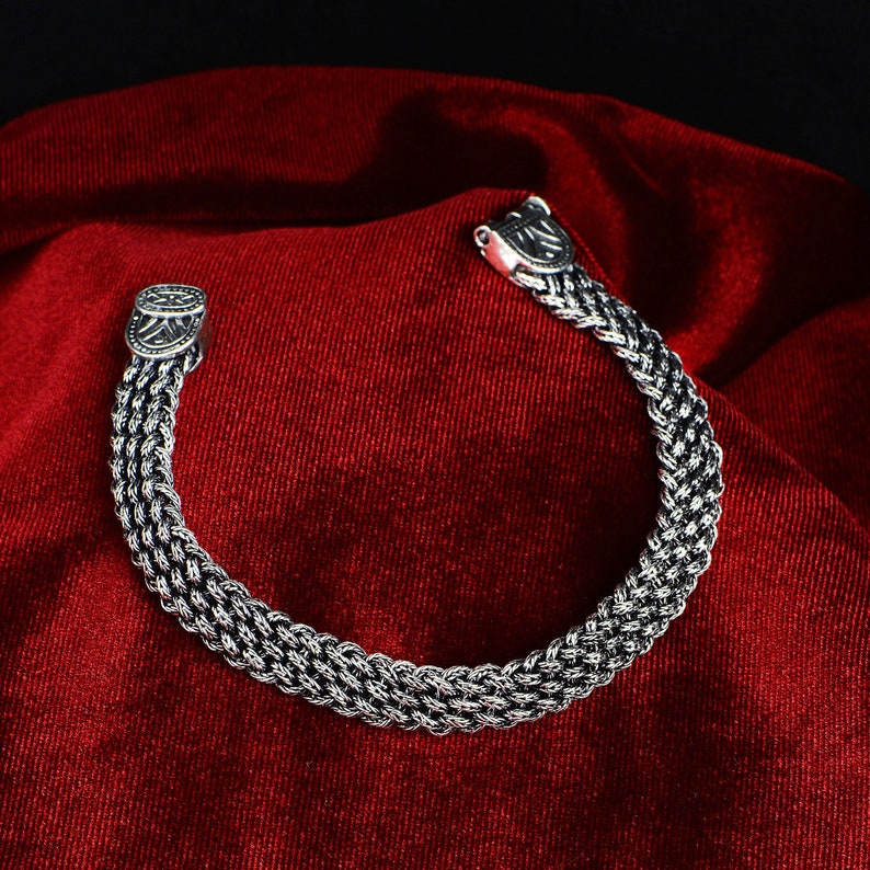 May include: A silver bracelet with a braided chain design and a clasp closure. The bracelet is on a red velvet background.