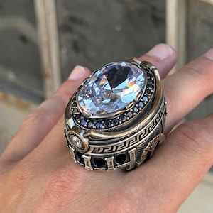 Puede incluir: Un gran anillo de plata adornado con una gran piedra preciosa ovalada y transparente como pieza central. El anillo tiene un diseño detallado de estilo antiguo con piedras preciosas negras más pequeñas y un patrón de greca alrededor de la banda.