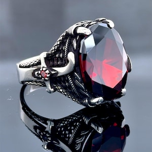 Puede incluir: Un anillo de plata con una gran piedra preciosa roja engastada en una garra. El anillo presenta diseños intrincados y detallados alrededor de la piedra preciosa.