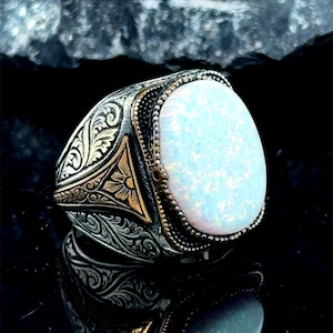 May include: A silver ring with an ornate design and a large white opal gemstone. The ring has a gold accent on the side.