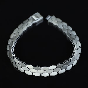Puede incluir: Una pulsera de tono plateado compuesta por eslabones metálicos hexagonales entrelazados. La pulsera presenta un acabado mate cepillado en la superficie de cada hexágono, con bordes oscuros y hundidos entre los segmentos y un cierre rectangular.