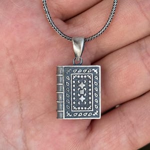May include: A silver pendant necklace featuring a detailed miniature book charm. The book charm has intricate patterns and a textured spine. The necklace chain is a woven design. The pendant is approximately 2.5 cm in height.