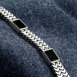 Puede incluir: Una pulsera plateada con un diseño de cadena trenzada y dos piedras rectangulares de ónix negro. La pulsera se muestra sobre un fondo de mezclilla azul, resaltando su brillo metálico y detalles oscuros.