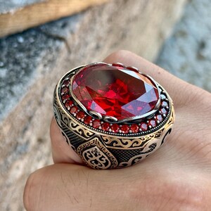 Puede incluir: Un anillo de plata ornamentado con una gran gema roja ovalada y facetada. El anillo presenta una banda grabada y est&aacute; rodeado de peque&ntilde;as piedras rojas. El dise&ntilde;o tiene una est&eacute;tica vintage. Los detalles sugieren artesan&iacute;a.