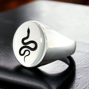 May include: A polished silver signet ring. The ring has a white oval face with a black snake design. The snake is stylized and coiled, adding a unique touch to the jewelry. The ring is a classic piece.