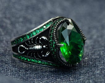 Puede incluir: Un anillo plateado con una gran piedra preciosa verde esmeralda facetada. El anillo presenta un diseño de escorpión en la banda y está adornado con piedras verdes más pequeñas. El anillo tiene un acabado oscuro y antiguo.