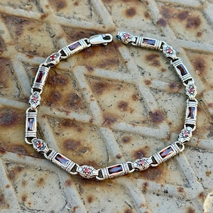 May include: Silver bracelet with rectangular red gemstones and small red accent stones. The bracelet has a decorative design with a clasp closure. The jewelry is displayed on a textured, metallic surface.